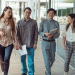Quatro trabalhadores caminham e conversam em um corredor de escritório moderno, carregando documentos.