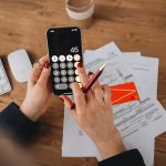 Mãos usando a calculadora de um smartphone sobre documentos financeiros.